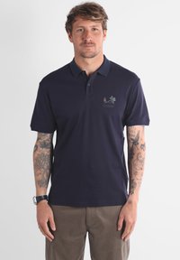 Polo shirt bleu marine à manches courtes, avec un col à trois boutons et un petit logo vert sur la poitrine. Fabriqué en tissu en coton lisse.