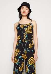 Macacão preto com padrão floral amarelo e detalhes em turquesa. Apresenta alças ajustáveis em espaguete e botões de madeira na parte da frente.