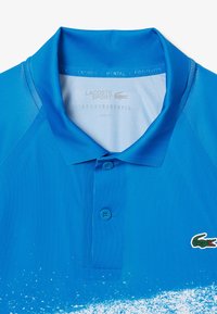 Sininen polo-paita, joka on valmistettu sileästä kankaasta. Paidassa on kaulus, kaksi nappia ja vihreä krokotiililogo. Gradientoituminen, jossa alaosa on vaaleampaa sinistä.