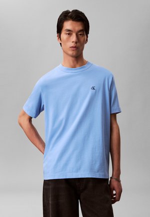 Mężczyzna w jasnoniebieskiej koszulce z krótkim rękawem z małym czarnym logo „cK” na piersi oraz ciemnych spodniach na szarym tle.