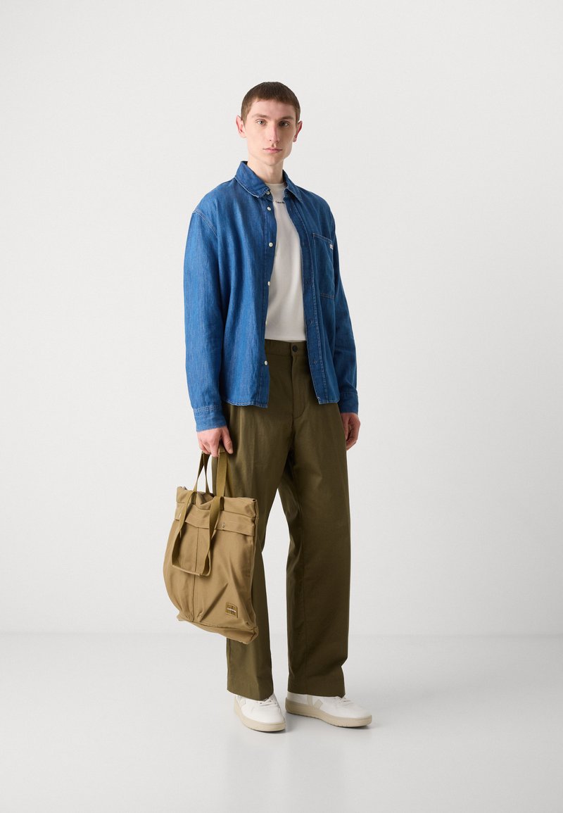 Denimhemd, weißes T-Shirt, olivgrüne Hose, beigefarbene Tasche mit Riemen und weiße Sneaker. Raum mit weißem Hintergrund, Modell steht aufrecht.