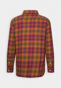 Chemise à manches longues boutonnée à carreaux dans des tons rouges, orange, verts et jaunes, vue de dos sur un fond uni.