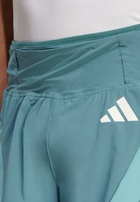 Primer plano de unos pantalones cortos deportivos en color teal con el logo blanco de Adidas y una cinturilla elástica, llevados sobre una tela de malla blanca.
