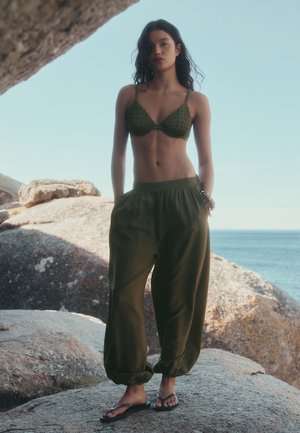 Jeune femme debout sur un rivage rocheux portant un bralette vert olive et un pantalon ample, avec l'océan et un ciel clair en arrière-plan.