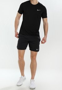 Camiseta negra de Nike de manga corta con un logo en el hombro izquierdo, combinada con pantalones cortos negros y zapatos deportivos blancos. Textura de tejido suave.