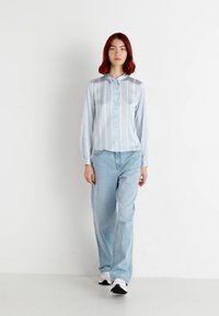 Camisa de botão azul claro de mangas longas com riscas verticais, combinada com jeans azul claro de corte largo e ténis. Textura suave.