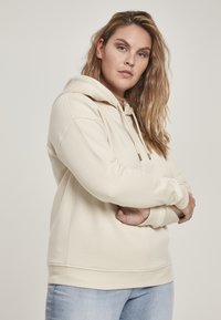 Bežová mikina vyrobená z mäkkého materiálu s predným vreckom na klokanie, sťahovacou kapucňou a rebrovanými manžetami. V kombinácii s svetlomodrými džínsami.