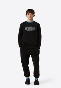 Sudadera negra con el logo de "The North Face" y detalles en azul, combinada con pantalones deportivos negros. El modelo lleva calzado negro y tobillos con calcetines azules.