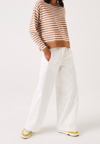 Person som bär en brun och vit randig tröja, vida vita byxor och vita sneakers med gula accenter, står med en hand i fickan.