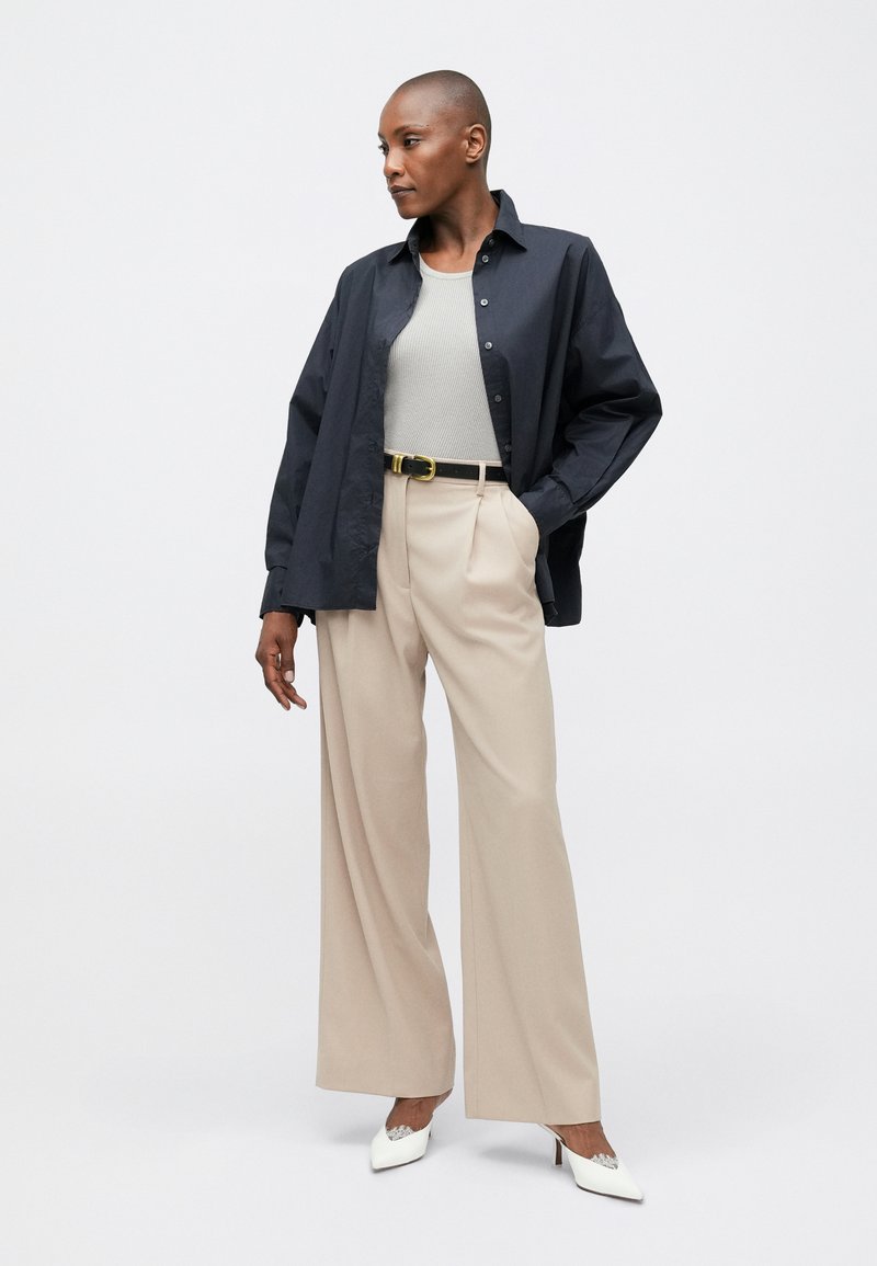 Camisa de botones azul oscuro, top de canalé gris claro, pantalones de alta cintura beige, cinturón negro con hebilla dorada, tacones blancos de punta.