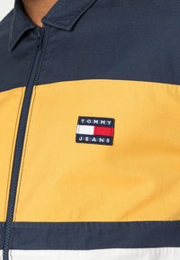 Marinblå och gul zip-up jacka med en strukturerad krage, som har en tygpatch-logotyp med "TOMMY JEANS" i blått, vitt och rött.