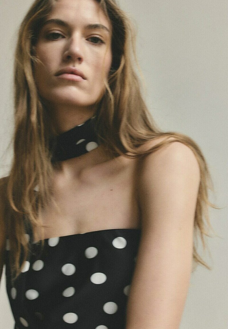 Jeune femme aux longs cheveux portant une robe noire bustier et un foulard assorti autour du cou avec de grands pois blancs sur un fond uni.