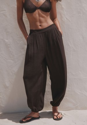 Femme portant un pantalon noir ample avec des poignets élastiques, un top noir transparent, des tongs et un collier en coquillage, debout contre un mur blanc texturé.