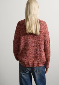 Suéter de punto en hilo rojo y multicolor con corte holgado, dobladillo acanalado y mangas largas. Combinado con vaqueros azul de tiro alto.