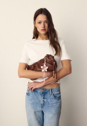 Jeune femme portant un haut blanc à manches courtes et un jean bleu, tenant un sac à main marron plissé avec un porte-clés en forme de fleur rose sur un fond uni.