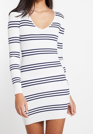 Kvinde iført en hvid ribbet langærmet kjole med horisontale mørkeblå striber og en dyb V-udskæring.