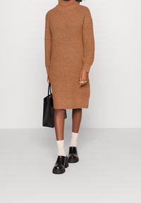 Abito a collo alto in maglia marrone, con maniche lunghe e taglio dritto. Indossato con scarpe nere con plateau e calze di colore chiaro. Borsa nera.