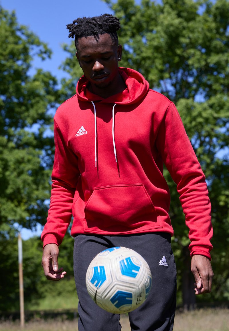 Roter Kapuzenpullover mit Kordelzügen und Fronttasche, kombiniert mit schwarzen Sporthosen mit Adidas-Logo. Ein Fußball wird kontrolliert.