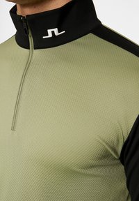 Grön och svart zip-up skjorta med hög krage, texturerad tyg och vitt logotyp på kragen, med kontrasterande färgpaneler.