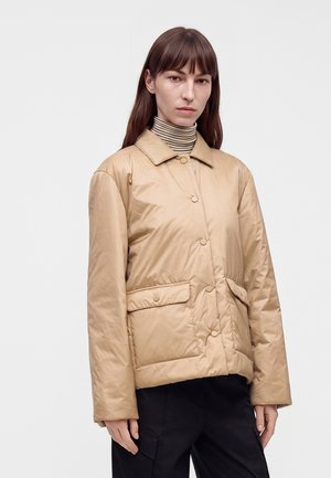 Femme aux cheveux bruns et lisses portant une veste matelassée beige avec de grandes poches et un col roulé rayé sur un fond uni.
