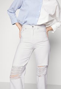 Calças de ganga brancas desgastadas com grandes recortes nos joelhos desfiados; design de cintura alta e textura suave. Combinadas com uma camisa riscada azul e branca.