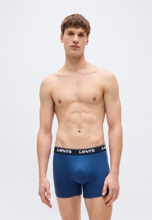 Jeune homme debout portant un boxer Levi's bleu avec une ceinture noire, fond clair uni, expression neutre, bras détendus.