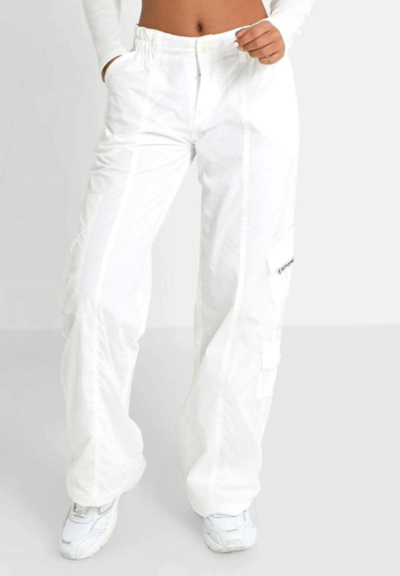 Femme portant un pantalon cargo blanc ample et des baskets blanches, avec une main dans la poche et un haut à manches longues raccourci laissant apparaître son ventre.