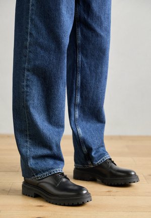 Schwarze Lederschuhe mit einer runden Zehenpartie, Schnürdesign und dicker, strukturierter Gummisohle, kombiniert mit lässig sitzenden blauen Jeans.