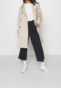 Beige trenčkot přes bílý tričko, doplněný námořnickými zkrácenými kalhotami a bílými vysokými teniskami. Obsahuje velké knoflíky a klasický design.