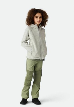 Barn med krøllet hår iført en lysegrå hættejakke med lynlås, grønne patchwork-bukser og sorte sko, stående med hænderne i lommerne.