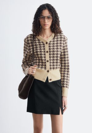 Femme aux cheveux bouclés portant de grandes lunettes, un cardigan à carreaux beige et marron, une jupe noire avec une fente, et tenant un sac à bandoulière marron.