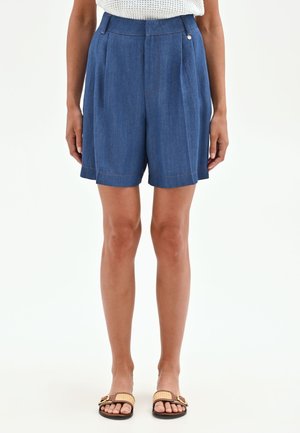 Femme portant un short en denim bleu taille haute avec des plis à l'avant et des sandales plates à brides tissées avec boucles dorées.