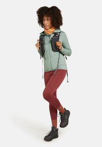 Tenue de randonnée comprenant une veste vert menthe, des leggings couleur rouille et des bottes de randonnée noires. Les accessoires incluent un cache-cou orange et un sac à dos noir.