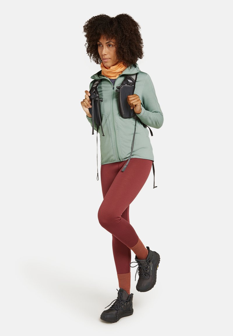 Tenue de randonnée comprenant une veste vert menthe, des leggings couleur rouille et des bottes de randonnée noires. Les accessoires incluent un cache-cou orange et un sac à dos noir.