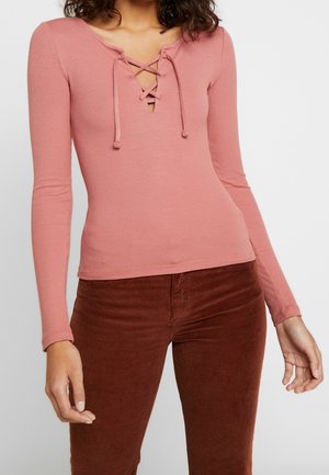 Vrouw draagt een aansluitende stofroze geribde top met lange mouwen en een gekruiste strikdetail bij de halslijn en een bruine corduroy broek.