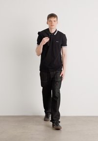 Crna polo majica s bijelim detaljima i malim logom, uparena s tamnim uzorkovanim hlačama. Model također drži crnu jaknu.