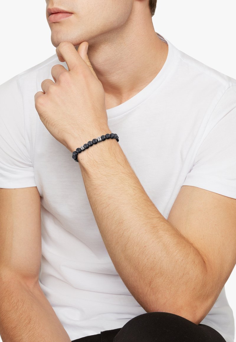 Svart pärlarmband med matt svarta och silverfärgade detaljer, som har runda pärlor på ett flexibelt band, bärs på ett handled mot en vit skjorta.