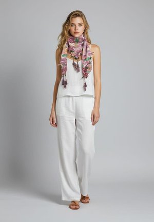 Joven mujer con camiseta blanca sin mangas y pantalones anchos, con una bufanda morada con flores y sandalias marrones, de pie frente a un fondo gris liso.
