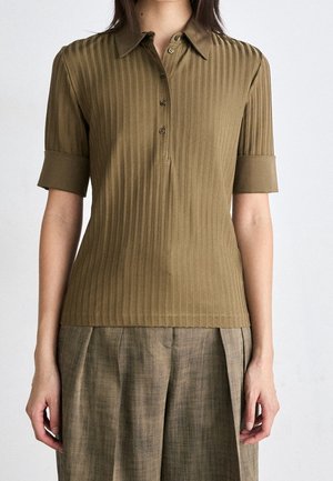 Kobieta ubrana w oliwkowo-zieloną prążkowaną koszulkę polo z krótkim rękawem z podwiniętymi mankietami oraz beżowe plisowane spodnie o szerokich nogawkach.