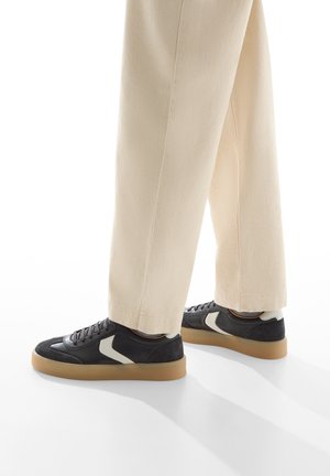 Sneakers in pelle nera con un design texture, dettagli bianchi e una suola in gomma color caramello, abbinati a pantaloni di colore crema.