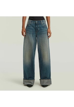 Wijde, vervaagde blauwe spijkerbroek met grote omslagen, gedragen door een persoon die staat tegen een effen lichte achtergrond.