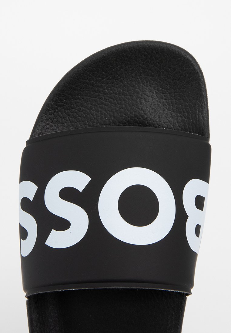 Sandale noire à glissière avec une sangle supérieure texturée ornée de grandes lettres blanches. Semelle intérieure et semelle douces pour plus de confort et de facilité à porter.