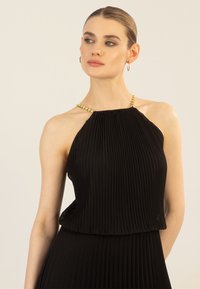 Musta laskostettu mekko, jossa on kaarroke-muotoilu ja monimutkainen kultaketju yksityiskohtana kaula-aukossa. Sujuva kangas lisää tyyliä ja eleganssia.