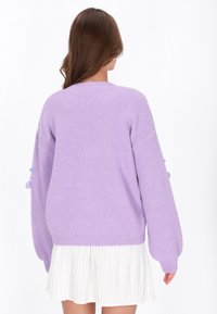 Fioletowy sweter żebrowany z oversize'owymi rękawami, z niebieskimi pomponami. Noszony na białej plisowanej spódnicy. Miękki, teksturowany materiał.