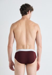 Burgundy férfi alsónadrág, fehér Calvin Klein logós derékszíjjal. Az anyag simának tűnik, szorosan illeszkedik, minták vagy egyéb részletek nélkül.