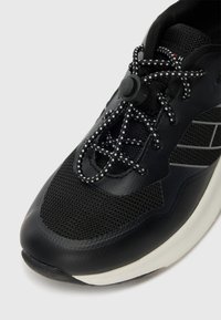 Basket de sport noire avec tige en mesh, semelle en caoutchouc et lacets élastiques. Présente une finition texturée et un bout arrondi. Rayures d'accent sur le côté.