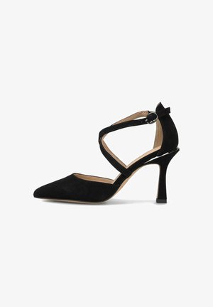 Zapato de tacón de ante negro con punta afilada, delgada correa al tobillo y tacón de aguja. Presenta una textura suave y un acento de hebilla circular.