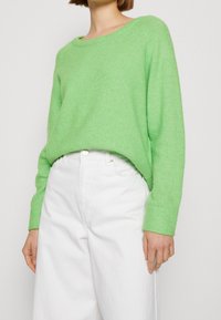 Personne portant un pull à manches longues vert vif rentré dans un pantalon blanc taille haute, mains détendues sur les côtés, fond neutre.