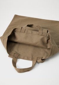Khaki värvi kangast ostukott, millel on kaks lühikest käepidet. Sisemus on voodrita ja avar, näidates tekstureeritud kangast viimistlust.