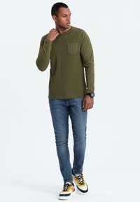 Top de manga larga en color verde oliva con canalé y un bolsillo en el pecho, combinado con jeans ajustados azules y zapatillas multicolores que presentan acentos en negro, amarillo y blanco.